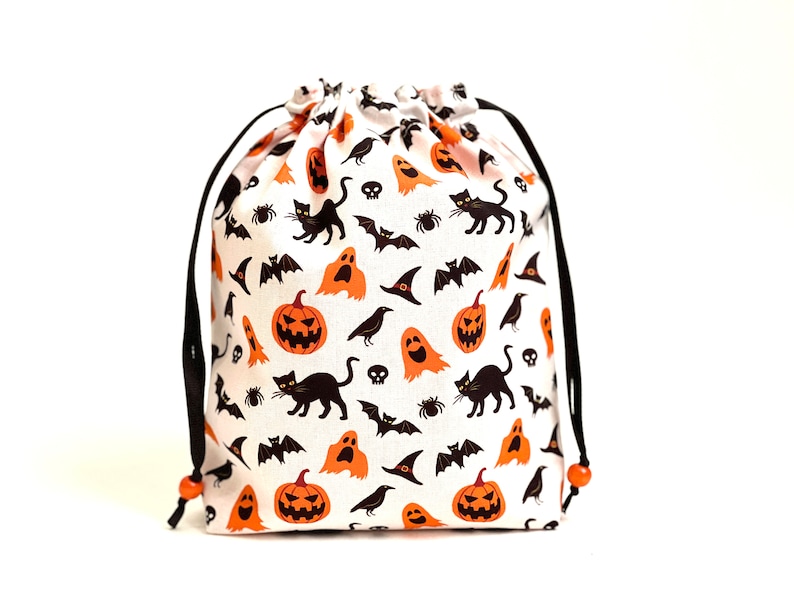 May include: A white drawstring bag with a Halloween print featuring orange pumpkins, black cats, bats, ghosts, and crows. The bag has black drawstrings with orange beads.