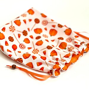 May include: A white drawstring bag with an orange and white print of pumpkins, candy corn, and lollipops. The bag has a brown wooden bead on the drawstring.