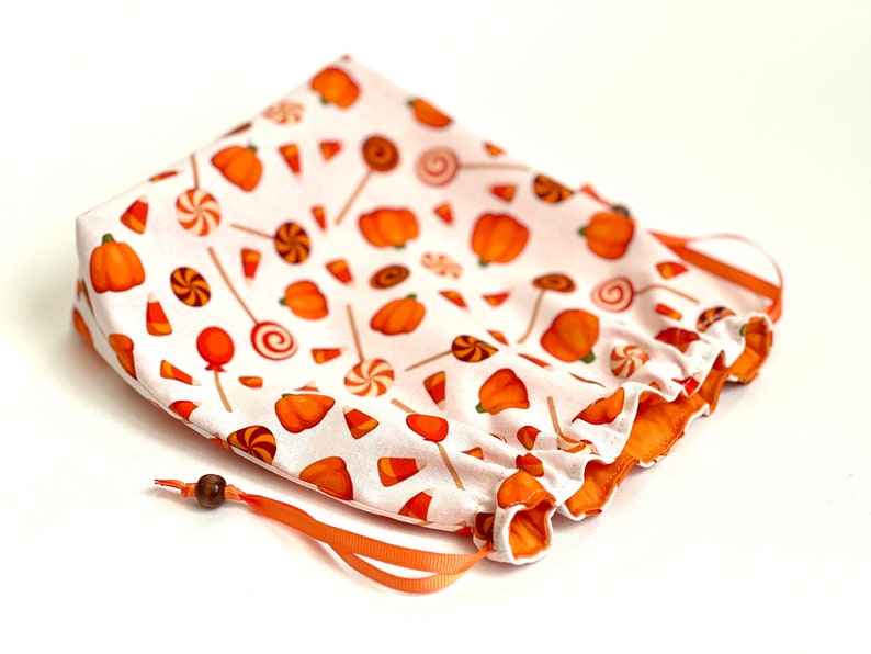 May include: A white drawstring bag with an orange and white print of pumpkins, candy corn, and lollipops. The bag has a brown wooden bead on the drawstring.