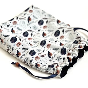May include: A white drawstring bag with a Halloween-themed print featuring bats, pumpkins, spiders, and the words "Boo!" and "Halloween". The bag has a navy blue drawstring and a wooden toggle.