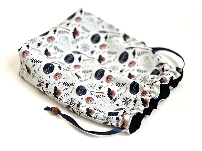 May include: A white drawstring bag with a Halloween-themed print featuring bats, pumpkins, spiders, and the words "Boo!" and "Halloween". The bag has a navy blue drawstring and a wooden toggle.