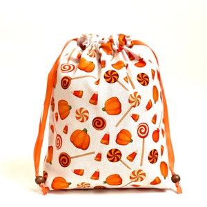 May include: A white drawstring bag with orange and white fabric featuring a pattern of pumpkins, candy corn, and lollipops. The bag has orange drawstrings with wooden toggles.