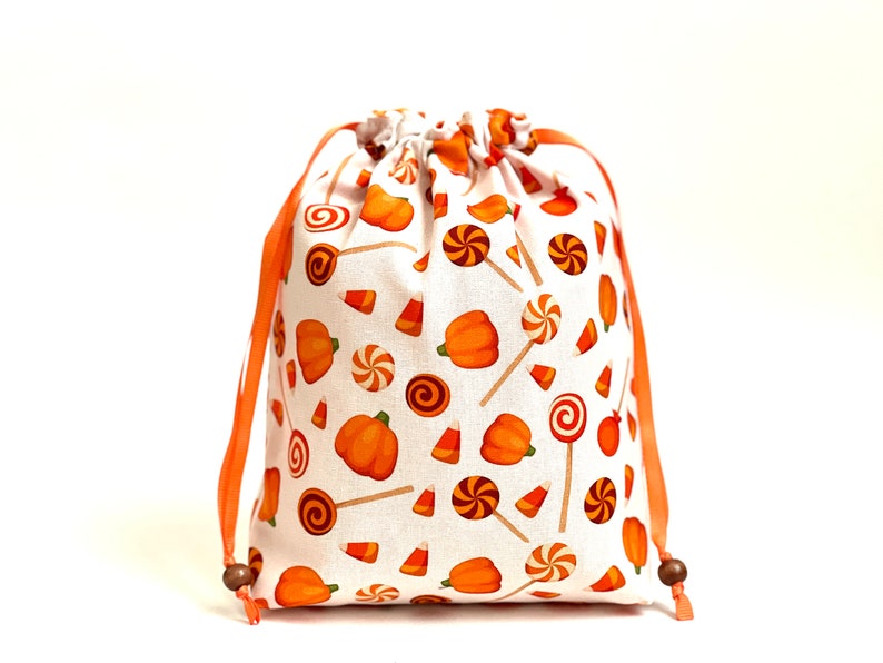 May include: A white drawstring bag with orange and white fabric featuring a pattern of pumpkins, candy corn, and lollipops. The bag has orange drawstrings with wooden toggles.