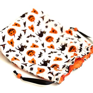 May include: A white drawstring bag with a Halloween-themed print. The print features orange pumpkins, black cats, bats, and ghosts. The bag has a black drawstring and orange lining.