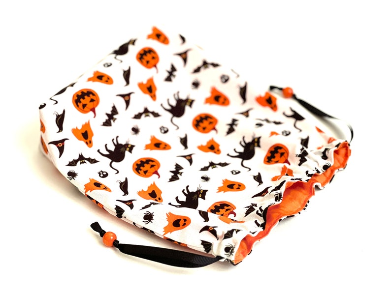May include: A white drawstring bag with a Halloween-themed print. The print features orange pumpkins, black cats, bats, and ghosts. The bag has a black drawstring and orange lining.