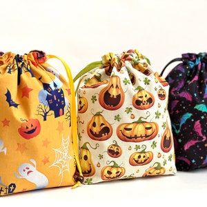 Op de afbeelding: Drie Halloween-thema trekkoordtassen. De eerste tas is geel met een spook, pompoen en ster patroon. De tweede tas is wit met een pompoen patroon. De derde tas is zwart met een vleermuis patroon.