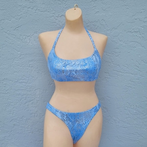 May include: A two-piece swimsuit set in a light blue snakeskin pattern. The top is a halter style, and the bottoms are a classic bikini cut. The swimsuit is displayed on a mannequin against a light blue textured wall.
