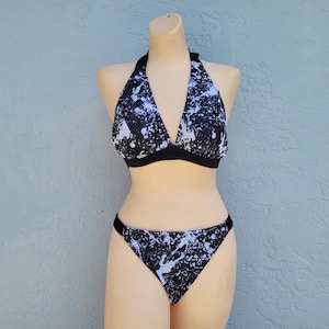 May include: A two-piece swimsuit set on a mannequin. The halter top and bikini bottoms feature a black and white splatter pattern. The edges of the top and bottoms are trimmed with black fabric. The background is a light blue wall.