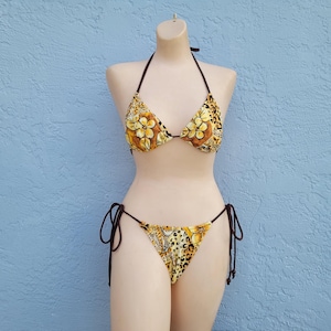 May include: A two-piece bikini set with a floral and animal print design. The top is a halter style with brown ties, and the bottoms are a string style. The bikini is displayed on a mannequin against a blue textured wall.