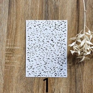 K&ouml;nnte beinhalten: Ein wei&szlig;es Blatt Papier mit einer schwarzen Silhouette von vielen V&ouml;geln darauf gedruckt. Das Papier liegt auf einer Holzoberfl&auml;che.
