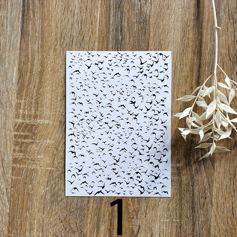 K&ouml;nnte beinhalten: Ein wei&szlig;es Blatt Papier mit einer schwarzen Silhouette von vielen V&ouml;geln darauf gedruckt. Das Papier liegt auf einer Holzoberfl&auml;che.
