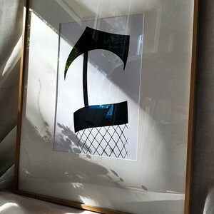 May include: A framed black and white print of a stylized letter 'T' with a grid pattern underneath. The print is in a light wood frame.