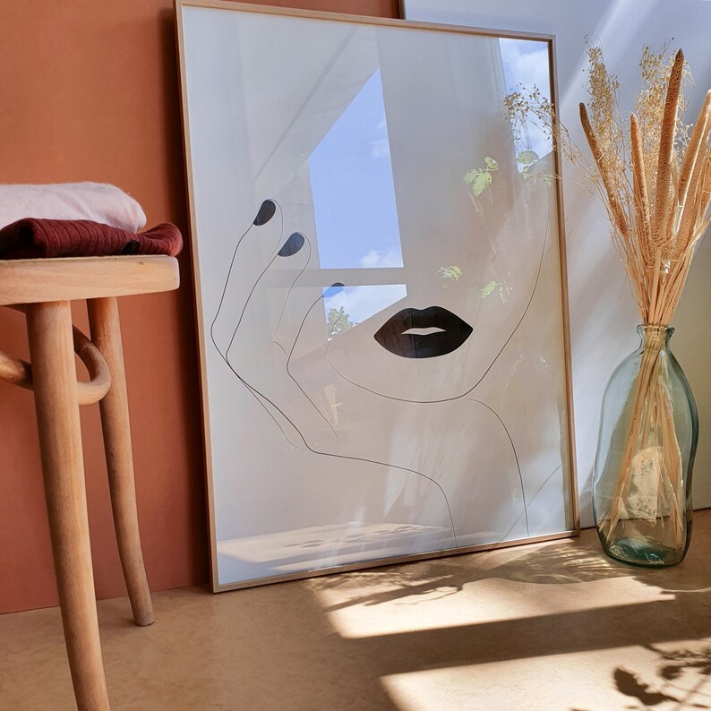 May include: A framed black and white line drawing of a woman's face with a closed eye, a hand, and a large, bold, black lip. The background is a blurred image of a window and sky.