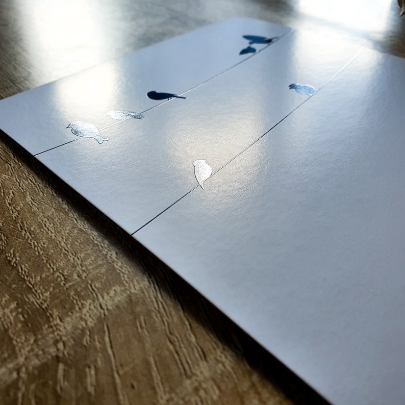 K&ouml;nnte beinhalten: Eine wei&szlig;e Karte mit einem silbernen Design von V&ouml;geln auf einem Draht. Die Karte liegt auf einer Holzoberfl&auml;che.