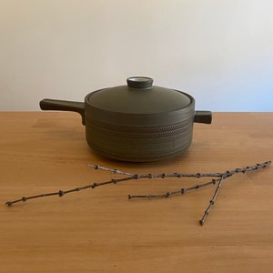 May include: A green ceramic pot with a lid and a textured design. The pot has a handle on each side. The pot is sitting on a wooden surface with a twig on the surface.