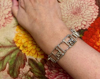 Könnte beinhalten: Ein silbernes Armband mit einem sich wiederholenden Muster aus kleinen, rechteckigen Rahmen, in denen sich jeweils eine stilisierte Tierfigur befindet.