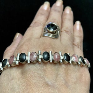 May include: A silver bracelet with alternating black and pink oval stones. The bracelet is made of linked sections with a silver bar between each stone. The bracelet is on a person's wrist.