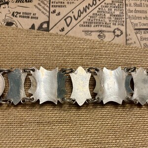 May include: A vintage silver-tone bracelet with a series of shield-shaped links connected by small rings. A U.S. quarter is placed next to the bracelet for size comparison. The bracelet is displayed on a tan fabric surface.
