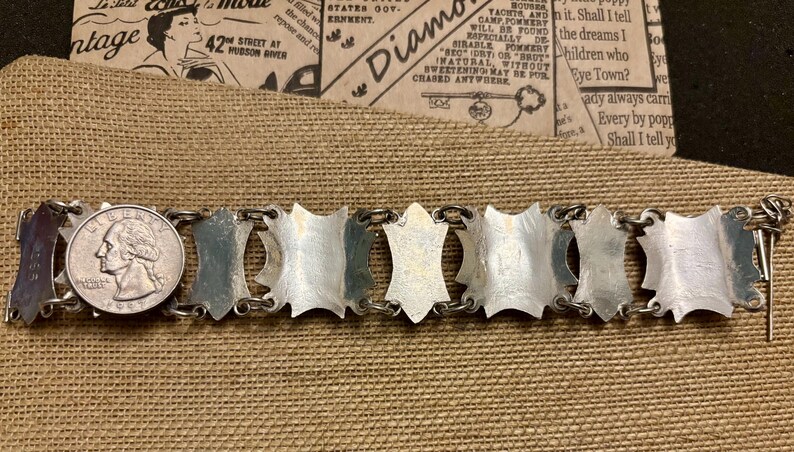 May include: A vintage silver-tone bracelet with a series of shield-shaped links connected by small rings. A U.S. quarter is placed next to the bracelet for size comparison. The bracelet is displayed on a tan fabric surface.