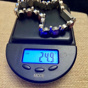 May include: A silver bracelet with a chain link design and round, multi-colored gemstones. The bracelet is on a digital scale that reads 24.9 grams.