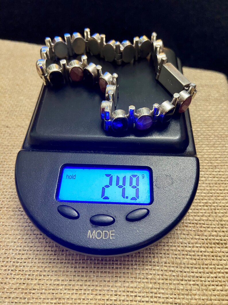 May include: A silver bracelet with a chain link design and round, multi-colored gemstones. The bracelet is on a digital scale that reads 24.9 grams.