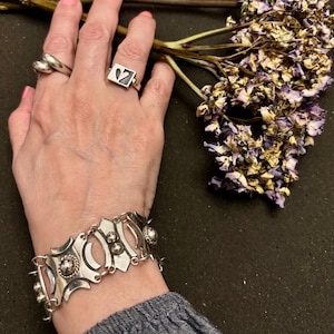 May include: A silver bracelet with an ornate design featuring oval and circular cutouts, and a matching ring with a heart-shaped design. The image also includes a second ring and dried purple flowers.