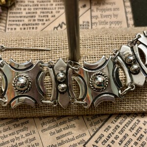 May include: A silver-toned bracelet with an ornate design. The bracelet features geometric shapes, textured details, and a secure clasp. The jewelry is displayed on a burlap-like surface.