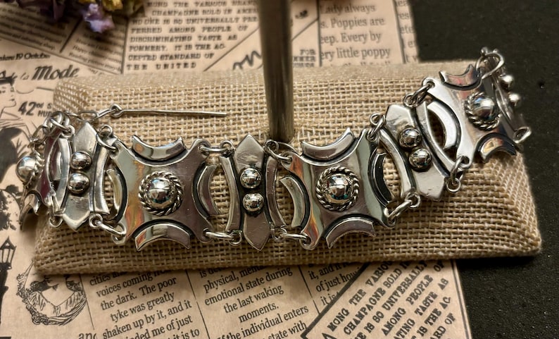 May include: A silver-toned bracelet with an ornate design. The bracelet features geometric shapes, textured details, and a secure clasp. The jewelry is displayed on a burlap-like surface.