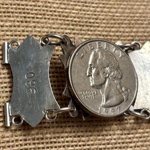 May include: Close-up of a silver bracelet with a quarter coin from 1997. The bracelet has a series of decorative shield-shaped links and circular links. The coin is placed in the center of the bracelet.