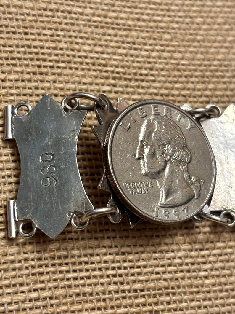 May include: Close-up of a silver bracelet with a quarter coin from 1997. The bracelet has a series of decorative shield-shaped links and circular links. The coin is placed in the center of the bracelet.