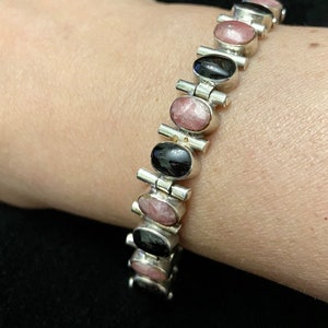 May include: A silver bracelet with alternating black and pink oval stones. The bracelet is made of linked metal pieces with a clasp.