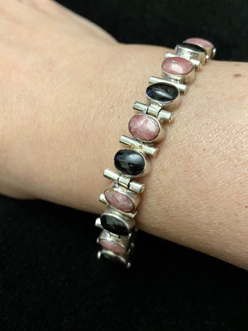May include: A silver bracelet with alternating black and pink oval stones. The bracelet is made of linked metal pieces with a clasp.