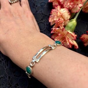 May include: Silver bracelet featuring oval green gemstones and a double-bar design. The bracelet is worn on a wrist, with a silver ring with a pearl-like center on a finger. Carnation flowers with red and yellow petals are in the background.