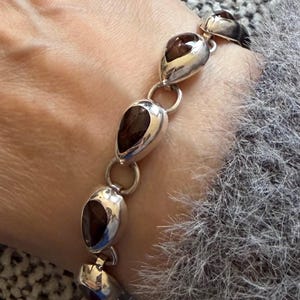 May include: A silver bracelet featuring teardrop-shaped, dark brown gemstones. The gemstones are set in polished silver settings and connected by small silver rings. The bracelet is shown on a wrist.