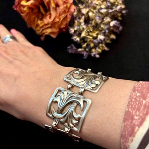 May include: Silver-toned bracelet featuring an ornate, openwork design. The bracelet is composed of rectangular links, each showcasing a stylized floral or foliate pattern. The links are connected by small, hinged connectors. The bracelet is worn on a wrist.