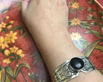 May include: A silver bracelet with an ornate design and a black onyx stone. The bracelet is on a person's wrist.