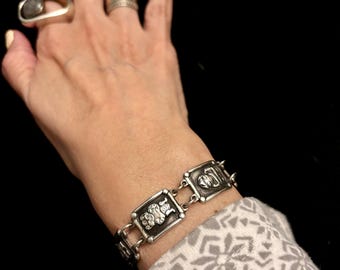 Könnte beinhalten: Silberfarbenes Armband mit rechteckigen Gliedern, die jeweils ein detailliertes geprägtes Design aufweisen. Das Armband wird am Handgelenk getragen, mit zwei Ringen an der Hand. Der Hintergrund ist schwarz.