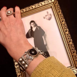 May include: A silver bracelet with a large, dark, oval stone and decorative metalwork is displayed. The bracelet is on a wrist, next to a framed black and white photograph. A silver ring with a circular design is also visible.