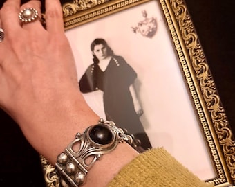 Könnte beinhalten: Ein silbernes Armband mit einem großen, dunklen, ovalen Stein und dekorativen Metallarbeiten wird gezeigt. Das Armband befindet sich an einem Handgelenk, neben einem gerahmten Schwarz-Weiß-Foto. Ein silberner Ring mit einem kreisförmigen Design ist ebenfalls sichtbar.