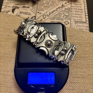May include: A silver-toned bracelet with geometric shapes and decorative accents rests on a digital scale. The scale displays a weight of 40.1, with the word "MODE" visible. The background includes vintage ephemera and dried flowers.