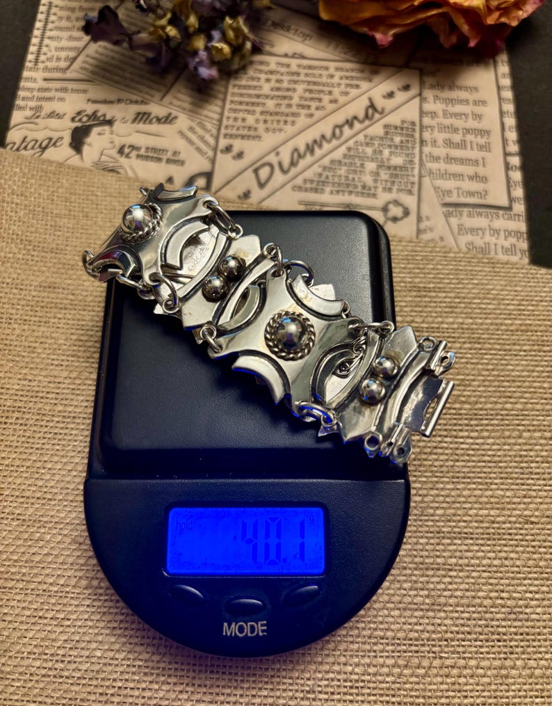 May include: A silver-toned bracelet with geometric shapes and decorative accents rests on a digital scale. The scale displays a weight of 40.1, with the word "MODE" visible. The background includes vintage ephemera and dried flowers.