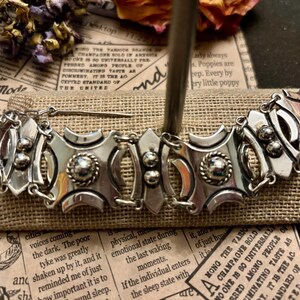 May include: A silver-toned metal bracelet with an ornate design. The bracelet features geometric shapes, circular accents, and a central decorative element. The piece is displayed on a burlap-textured surface.
