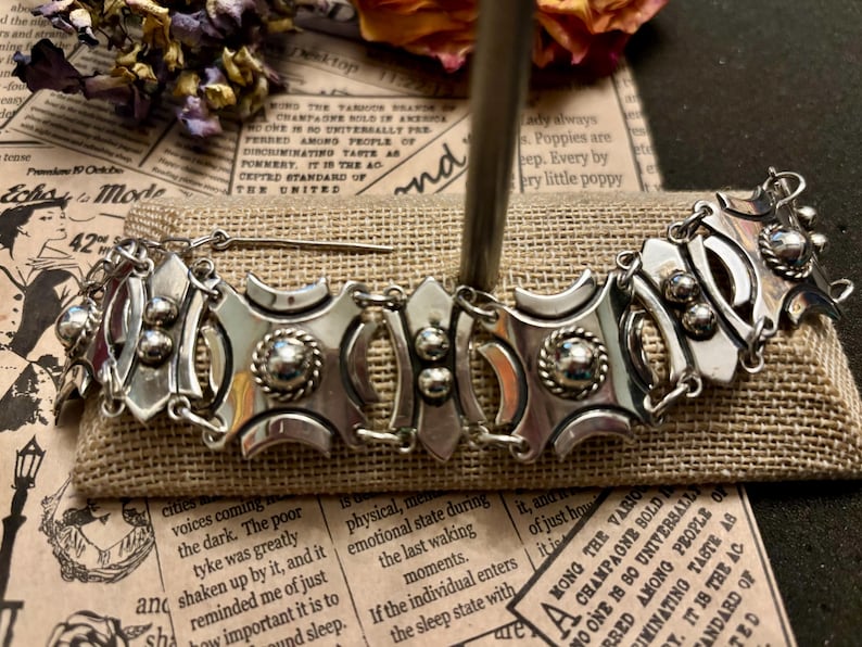 May include: A silver-toned metal bracelet with an ornate design. The bracelet features geometric shapes, circular accents, and a central decorative element. The piece is displayed on a burlap-textured surface.