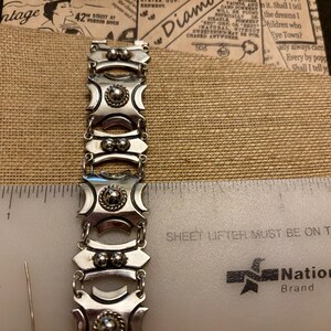 May include: A silver-toned bracelet with a geometric design, featuring alternating rectangular and oval links. Each link has a central raised circular element. The bracelet is measured with a ruler in inches.