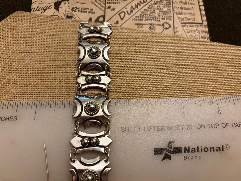May include: A silver-toned bracelet with a geometric design, featuring alternating rectangular and oval links. Each link has a central raised circular element. The bracelet is measured with a ruler in inches.