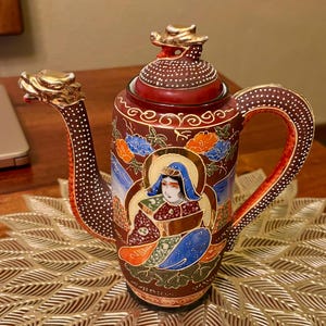 May include: An ornate, antique teapot with a dragon-shaped spout and handle. The teapot is primarily dark red with gold accents and a painted scene of a woman in a blue headdress. Floral and geometric patterns decorate the body and lid.