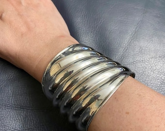 May include: A wide, polished silver cuff bracelet with a ribbed design. The bracelet is worn on a wrist, reflecting light. A silver ring with a decorative design is also visible.