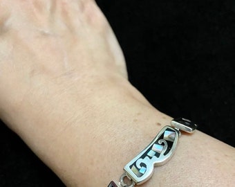 May include: A silver bracelet with black and white inlay. The bracelet is made of a series of connected links, each with a curved shape and a decorative design.