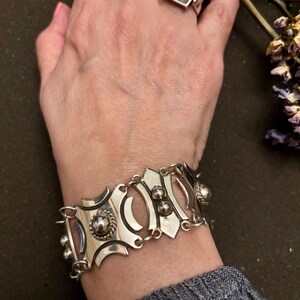 May include: A silver bracelet with an ornate design featuring circular and oval shapes, and small ball accents. The bracelet is connected by small links. Two silver rings are also visible.