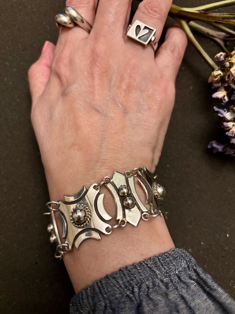May include: A silver bracelet with an ornate design featuring circular and oval shapes, and small ball accents. The bracelet is connected by small links. Two silver rings are also visible.